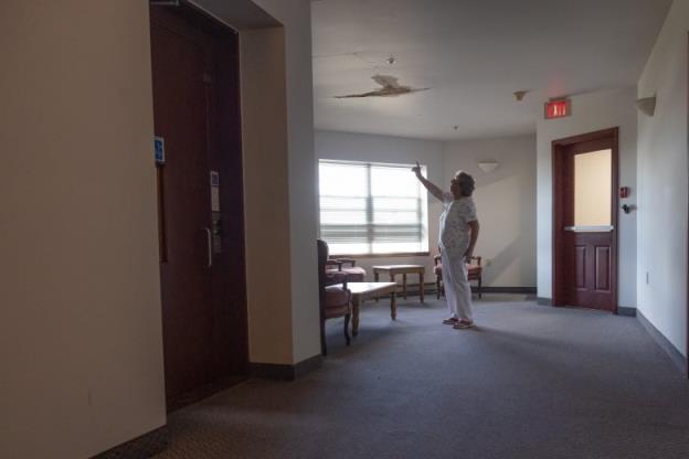 An elderly woman stands in a hallway and points at a large cracked area in the ceiling above. 